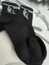 Par de medias blancas de algodón con puño acanalado con una línea negra, logo de la marca en el tobillo y estampado de alas negras.