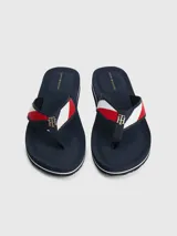 Ojotas de mujer color azul marino con tira de tela trenzada que presenta franjas rojas y blancas, y un dije metálico con el monograma TH en dorado.