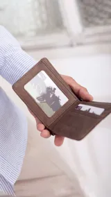 Billetera de cuero sintético color marrón oscuro con textura granulada, presenta una banda elástica decorativa central con rayas blancas y tostadas. Cuenta con compartimentos internos para billetes, tarjetas y visor.