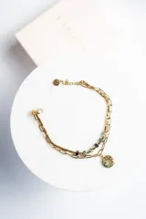 Pulsera dorada de acero quirúrgico con doble cadena, una de eslabones y otra con cuentas verdes y beige. Dije circular texturizado con piedra verde central.