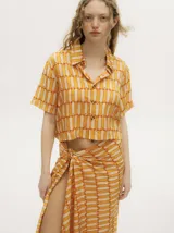 Camisa crop de manga corta con cuello camisero y cierre frontal de botones. Presenta un diseño geométrico estampado en tonos naranja y crema, con un calce suelto y liviano.