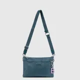 Mini cartera tipo bandolera de la marca Bubba, color azul petróleo, con correa ajustable de tela grabada con el logo de la marca. Incluye un dije colgante con la letra B en color lila y cierre superior.