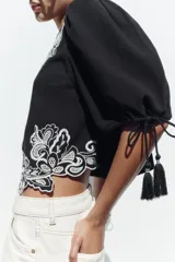 Blusa corta negra con mangas abullonadas y bordados florales en contraste de color blanco. Presenta cuello redondo con puntilla, mangas ajustables con cordones y borlas, y cierre en la espalda con abertura y botón oculto.
