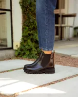 Bota tipo Chelsea de cuero negro con paneles elásticos laterales. Presenta una suela track de goma y un tirador de cuero marrón en la parte posterior.