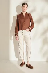 Blusa de lino color marrón con escote en V, cuello con volados y mangas largas con puños fruncidos que presentan detalles en color crudo, al igual que en los hombros. Cuenta con cierre frontal de botones.