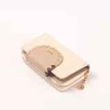Billetera color beige con cierre dorado y detalle de solapa decorativa en tono más oscuro con borde trenzado y logo dorado. Incluye un charm colgante con el logo de la marca.