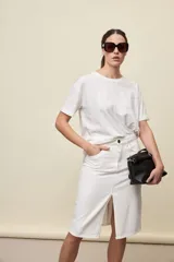 Falda midi de jean color blanco, de corte recto, con tiro alto, cinco bolsillos, cierre frontal con botón y abertura central en la parte delantera.
