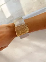 Brazalete ancho de textura plateada con un chapón central rectangular en tono dorado con relieve ondulado. Cuenta con cierre imantado.