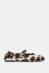 Zapato plano tipo bailarina con acabado de piel y estampado animal print de manchas marrones y blancas. Presenta una tira ajustable con hebilla metálica sobre el empeine y punta redonda.