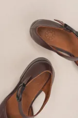 Zapato de cuero color borgoña, con puntera redondeada y diseño trenzado. Presenta una tira sobre el empeine con hebilla metálica negra.