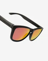 Lentes de sol Hawkers modelo One, con armazón negro mate y cristales polarizados con efecto espejado en tono rojizo.