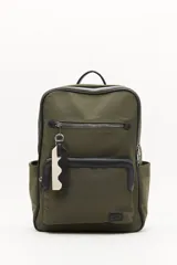 Mochila urbana de nylon en color verde militar, con detalles en cuero sintético negro. Cuenta con compartimento principal con cierre, bolsillo frontal con cierre, bolsillos laterales abiertos y asa superior. Incluye un colgante decorativo de tela y correas ajustables acolchadas.