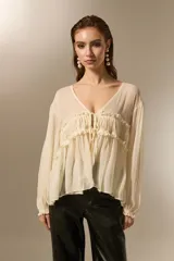 Blusa de poliéster color natural, con cuello en V, mangas largas con puños elásticos y detalle de frunces con cordón ajustable en el frente.