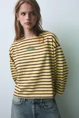 Camiseta oversize de manga larga con diseño de rayas horizontales amarillas y negras. Presenta cuello redondo y un detalle de texto bordado en el centro del pecho.
