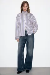 Camisa de manga larga con cuello subido y trabillas en los hombros. Presenta un diseño de rayas verticales en tonos azul, beige y marrón, con cierre frontal de botones.