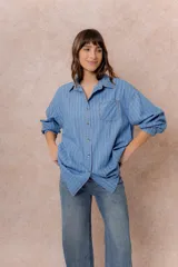 Camisa oversize de tela liviana con efecto denim, color azul con rayas verticales blancas. Presenta cuello camisero, cierre frontal con botones, bolsillo aplicado en el pecho, hombros caídos y mangas largas con puños abotonados.