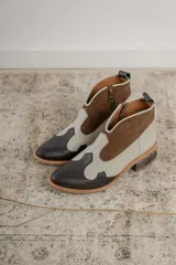 Botinetas estilo western de caña baja, confeccionadas en una combinación de cuero liso y gamuza en tonos marrón oscuro, marrón medio y crema. Presentan cierre lateral, taco bajo cuadrado y diseño con recortes característicos del estilo cowboy.