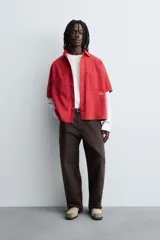 Camisa de algodón con estructura canvas, color rojo lavado, de corte relajado (relaxed fit) y manga corta. Posee cuello solapa, dos bolsillos de plastrón en el pecho y cierre frontal de botonadura. Presenta bordado y estampado de texto a contraste en el delantero.