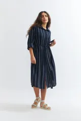 Vestido camisero midi de lino color azul marino con rayas verticales blancas, corte holgado, cuello camisero, mangas 3/4 con puños elásticos y lazo en la cintura.