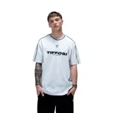 Remera unisex de color blanco con cuello en V, mangas cortas y detalles de líneas finas en color oscuro en los hombros y laterales. Presenta el logo de la marca estampado en el pecho junto con un pequeño parche decorativo.