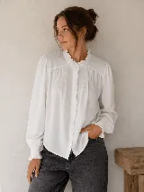 Camisa negra de manga larga con detalles de volados en el cuello alto, a lo largo de la botonadura frontal y en los puños elásticos. Presenta frunces en el canesú delantero y cierre frontal con botones.