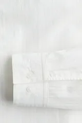 Camisa blanca de corte regular fit, confeccionada en una mezcla suave de lyocell y algodón. Presenta cuello inglés, tapeta clásica, bolsillo superior abierto en el pecho y canesú con pliegue de caja en la espalda. Mangas largas con abertura y puños ajustables con botón. El bajo es redondeado.