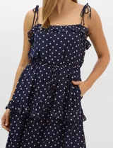 Vestido largo azul marino con estampado de lunares blancos, confeccionado en gasa fluida. Cuenta con tirantes finos ajustables con lazos en los hombros, escote con volados, corte en la cintura con lazo ajustable, y falda larga con múltiples capas y volados horizontales.