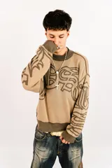 Sweater tejido reversible de corte boxy fit, con cuello redondo y puños acanalados. Presenta un diseño gráfico abstracto de estilo gótico que recorre el pecho y las mangas. Un lado es de color marrón oscuro con detalles en beige, y el otro lado es de color beige con detalles en marrón oscuro.