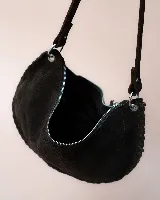 Bolso tipo hobo de gamuza color marrón oscuro, con diseño de media luna y detalles trenzados en los bordes. Cuenta con cierre superior, tiradores de cuero anudados y correa ajustable y desmontable con herrajes metálicos.