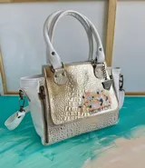 Cartera de cuero blanca y dorada con solapa texturizada tipo cocodrilo y detalles pintados a mano. Cuenta con doble asa de mano, correa ajustable, herrajes metálicos y cierre superior.