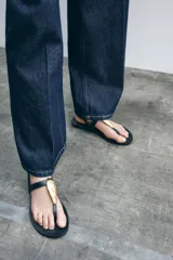 Sandalia plana negra con tira para el dedo y tira tobillera ajustable con hebilla dorada. Presenta un adorno grande de pieza metálica dorada con textura orgánica sobre el empeine.