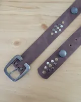 Cinto de cuero marrón con hebilla metálica de estilo envejecido y detalle de tachas metálicas en diferentes tonos en el extremo.
