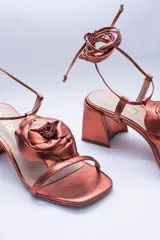 Sandalias de cuero color cobre con taco cuadrado y tira fina sobre los dedos. Presentan una flor grande en el empeine y tiras finas para atar al tobillo.