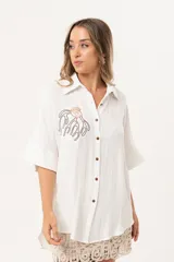 Camisa blanca de silueta oversize, con cuello camisero, manga 3/4 y corte más largo en la espalda. Presenta bordados de coral y caracol en el frente, y botones a tono.