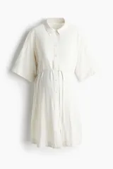 Vestido corto camisero de viscosa color blanco, con cuello camisero, botonadura frontal completa y mangas cortas con hombros caídos. Incluye un cinto fino ajustable debajo del busto.