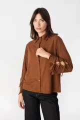Camisa de manga larga color camel con textura geométrica sutil. Presenta cuello clásico, cierre frontal con botones y detalle de tiras ajustables en las mangas.