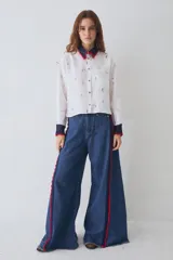 Camisa de lino blanca con estampado de motivos deportivos, cuello y puños tejidos a mano en crochet azul con borde rojo. Presenta cierre frontal con botones y un bolsillo en el pecho.
