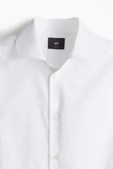 Camisa blanca de algodón con cuello italiano, tapeta clásica y canesú en la espalda. Mangas largas con tapeta y botón, y cierre ajustable de botón en los puños. Bajo levemente redondeado y corte ajustado.