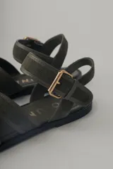 Sandalia plana de cuero negro, con diseño minimalista de tiras finas cruzadas sobre el empeine y una tira ajustable con hebilla metálica en el tobillo.
