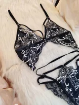 Conjunto de lencería compuesto por un bralette triangular y una tanga, confeccionados en encaje negro con detalles florales en color hueso. El bralette presenta breteles ajustables y diseño con tiras decorativas, mientras que la tanga cuenta con doble tira lateral regulable.