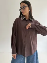 Camisa oversize de color marrón con cuello camisero y puños con detalle de bordado festoneado en color blanco. Presenta cierre frontal de botones y mangas largas.
