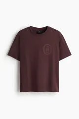 Remera de punto de algodón de peso medio color bordó, con cuello redondo ribeteado, hombros caídos y bajo recto. Presenta un diseño estampado circular en el pecho con texto y un arco. Corte holgado.