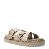 Sandalias planas Miss Carol modelo Zelv, color beige, con tiras anchas decoradas con hebillas metálicas doradas y tachas, sobre suela de plataforma baja con costuras visibles.