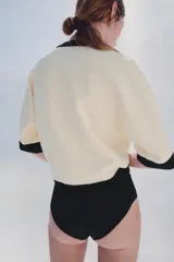 Chaqueta de punto color crudo con ribetes negros en el cuello en V, la botonera, el bajo y los puños de las mangas cortas. Cuenta con cierre frontal con botones dorados grabados.