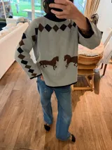 Sweater de tejido de punto en color gris claro, con diseño de caballos en color marrón en la parte frontal y patrón de rombos en los hombros y mangas. Presenta cuello, puños y ruedo en color azul marino, con un corte holgado y cómodo.