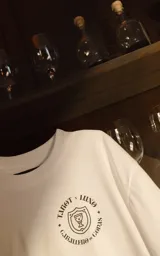 Remera blanca de corte oversize y manga corta, confeccionada en algodón premium. Presenta un estampado gráfico en la espalda con una ilustración estilo cómic de un caballero medieval con una copa y un celular, haciendo referencia a la carta del tarot 'Caballero de copas'.