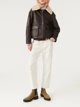 Chaqueta corta de cuero color marrón con cuello de piel sintética color crema, cierre con botones a presión, bolsillos frontales con solapa y puños con botón.