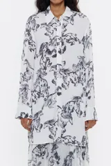 Camisa larga blanca con estampado de galgos negros, corte recto, cuello camisero, manga larga con puños abotonados y cierre frontal con botones.