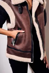Chaleco oversize estilo gamulán en color marrón, confeccionado en cuero vegano con forro y solapas de corderito sintético en contraste. Posee cierre frontal metálico, bolsillos con cremallera y detalles de correas con hebillas en el cuello.