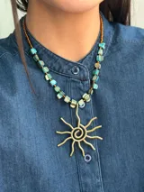 Collar corto con dije central en forma de sol, elaborado en bronce, con cadena compuesta por cuentas de nácar en tonos turquesa y mostacillas marrones.